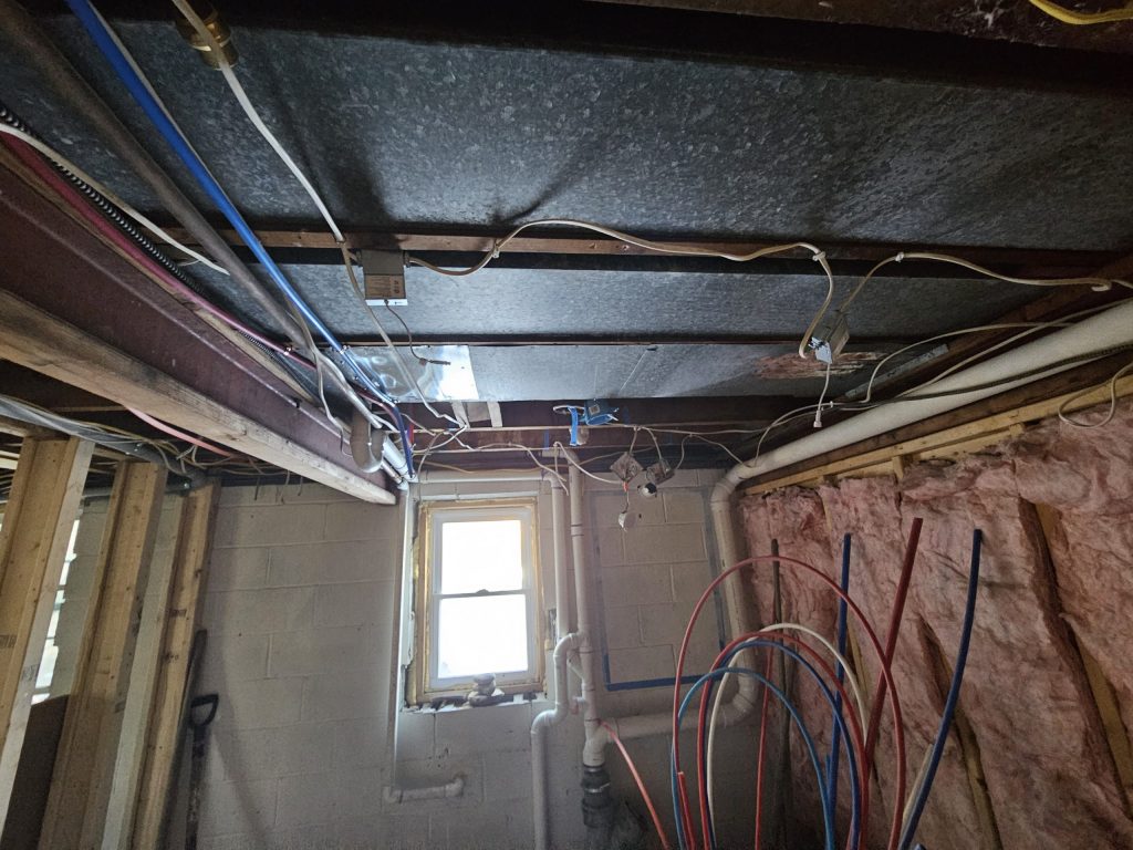 The bathroom corner before work: concrete block walls, a small window, exposed plumbing lines, pink insulation and overhead ductwork and wiring.