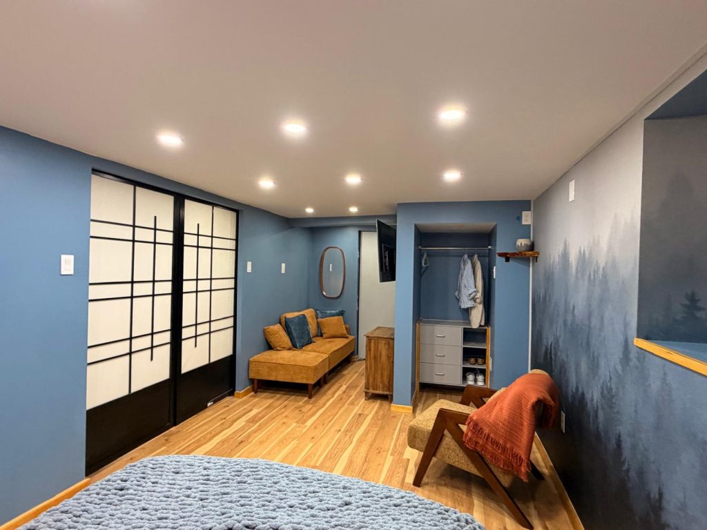 A restrained bedroom in dusty blue with black-and-white sliding doors, a built-in closet, oak floor and a forest mural on the far wall.