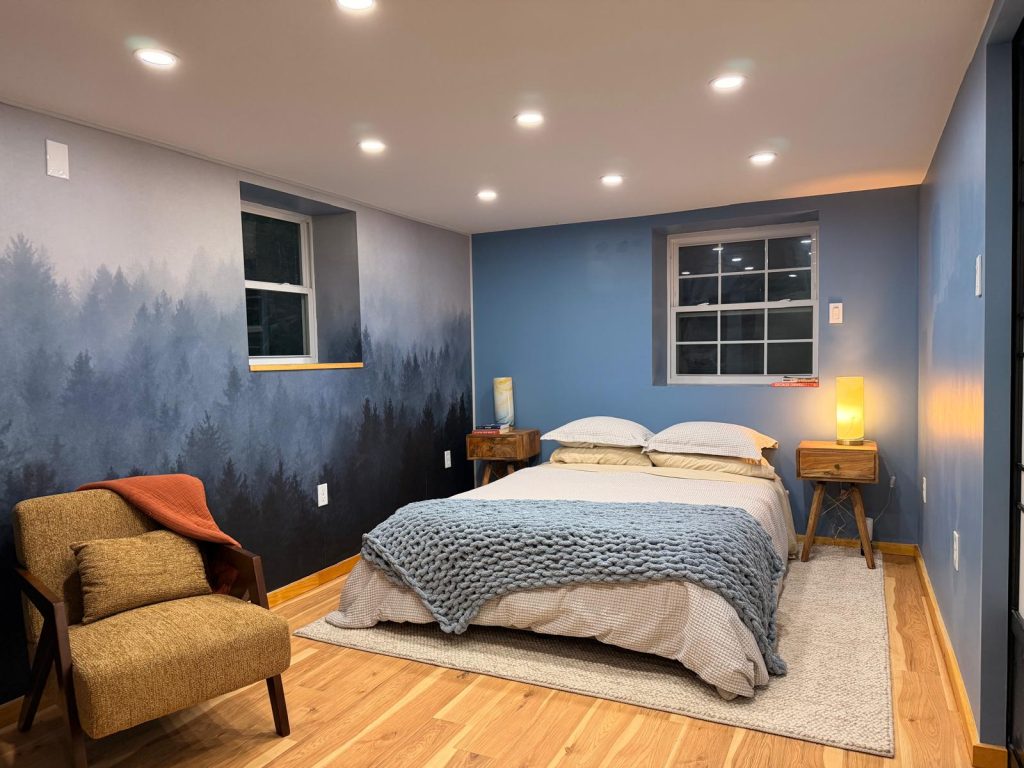 A finished bedroom with a misty forest mural wall, dusty blue paint, oak floor and a mustard mid-century armchair.