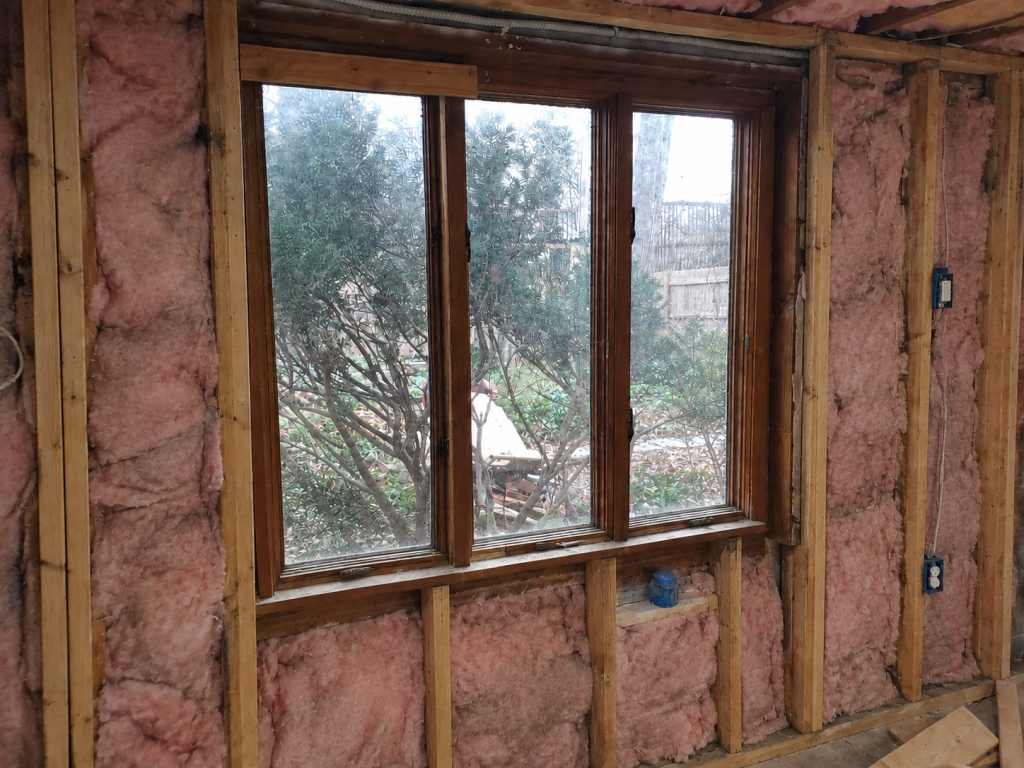 The bay window during construction: open stud walls packed with pink insulation, old dark-framed windows, bare wiring and outlet boxes.