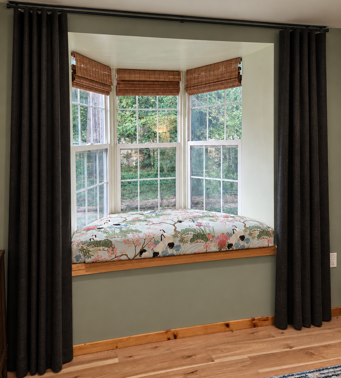 A deep bay window seat cushioned in Japanese crane-and-garden fabric, framed by bamboo shades and charcoal curtains, looking into a green garden.