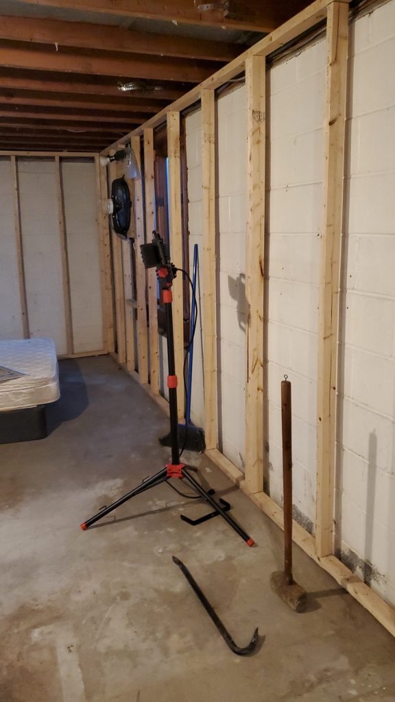 Looking down the passage during the build: new stud framing against painted block, bare concrete floor, a work light on a tripod and a sledgehammer leaning on the wall.
