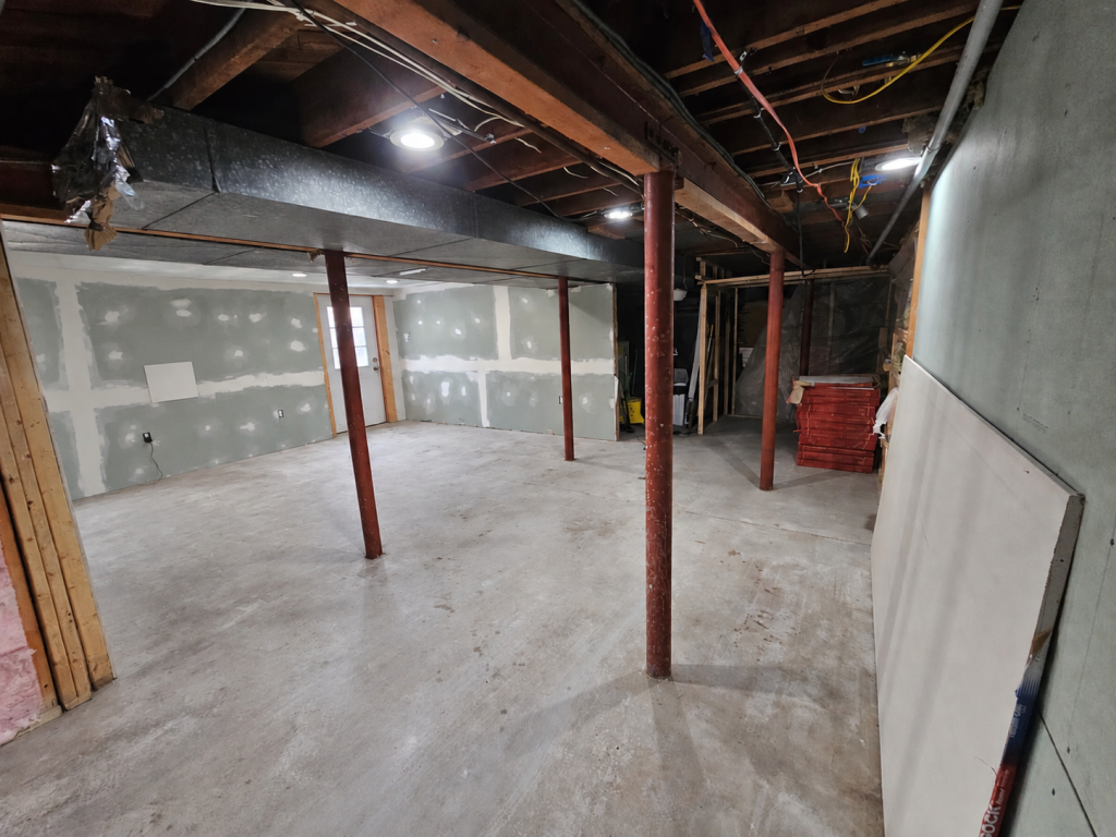 The kitchen area before work: bare concrete floor, red steel support columns, taped drywall and open joists with exposed wiring and ductwork.