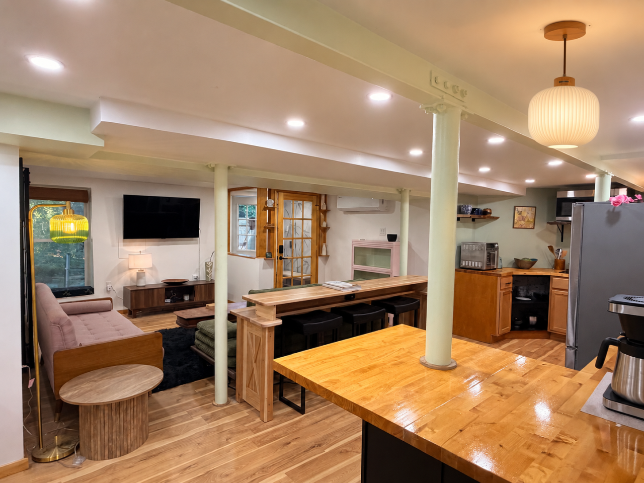 The finished open-plan kitchen and living room: oak floor, butcher-block counter, pale green wood-clad columns, a ribbed pendant lamp and a sitting area beyond.