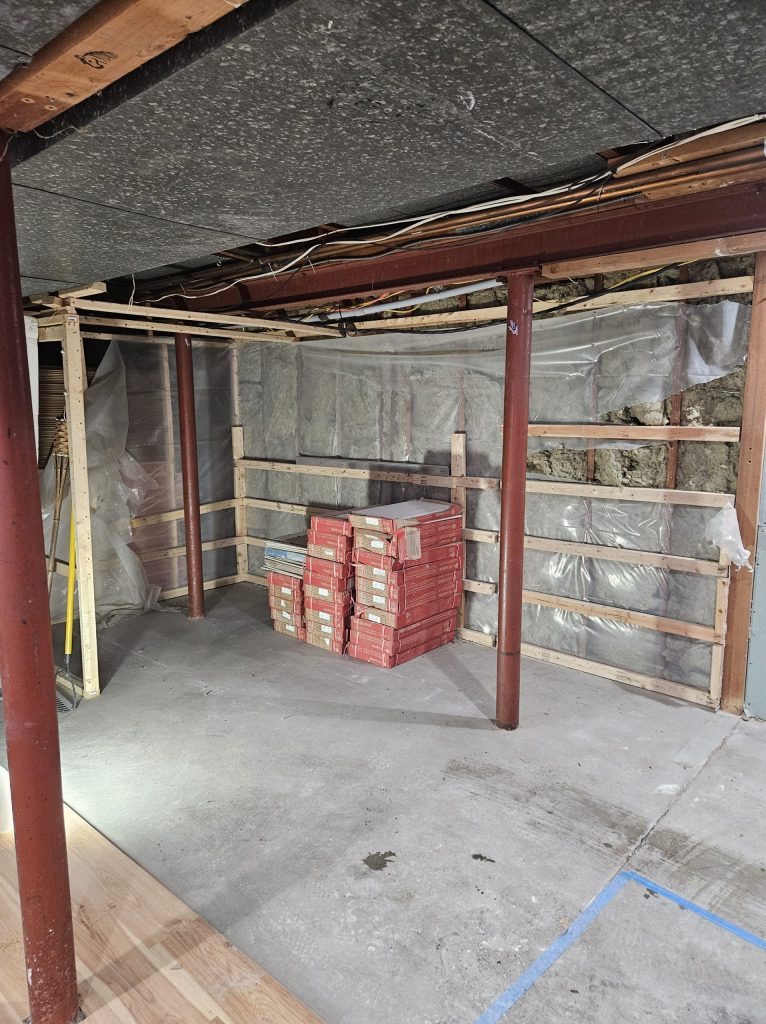 The space during framing: red steel columns, new stud walls, insulation behind plastic sheeting and boxes of flooring stacked on the concrete.