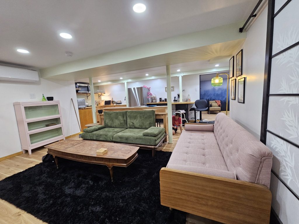 The finished living room with wood-clad columns, soft green walls, layered lighting and large windows onto the meadow.