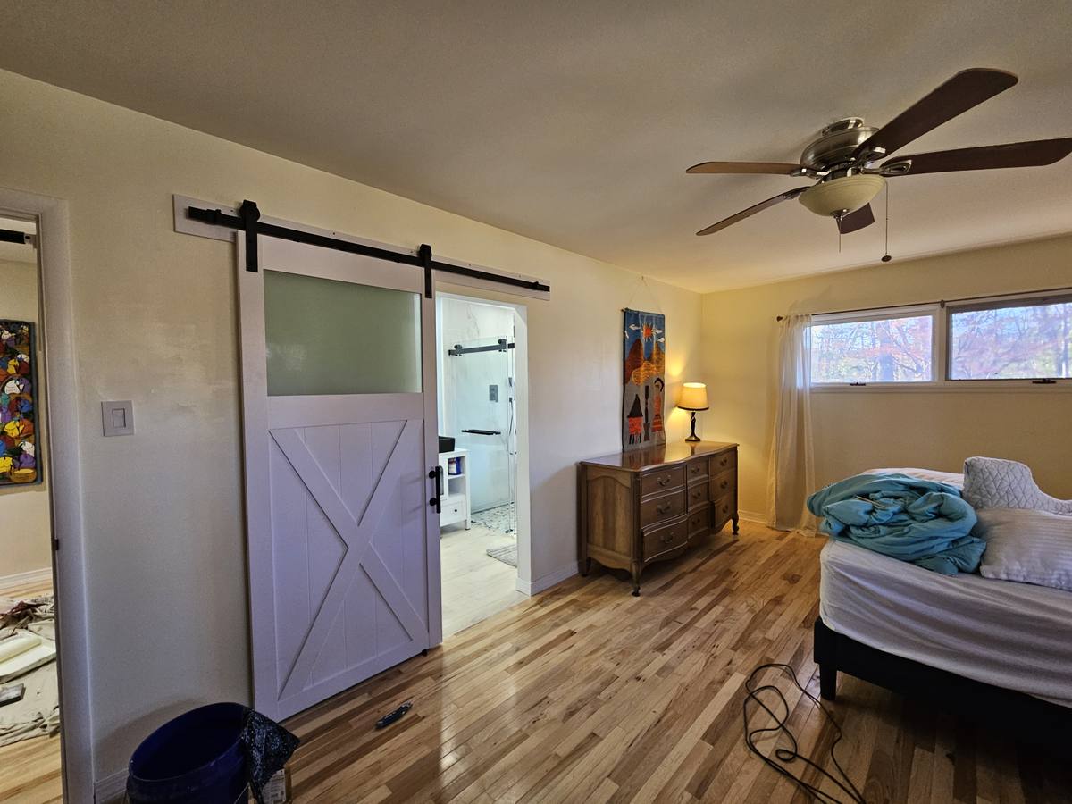 A bedroom with a sliding barn door standing open to the next room, and light from a corner window.