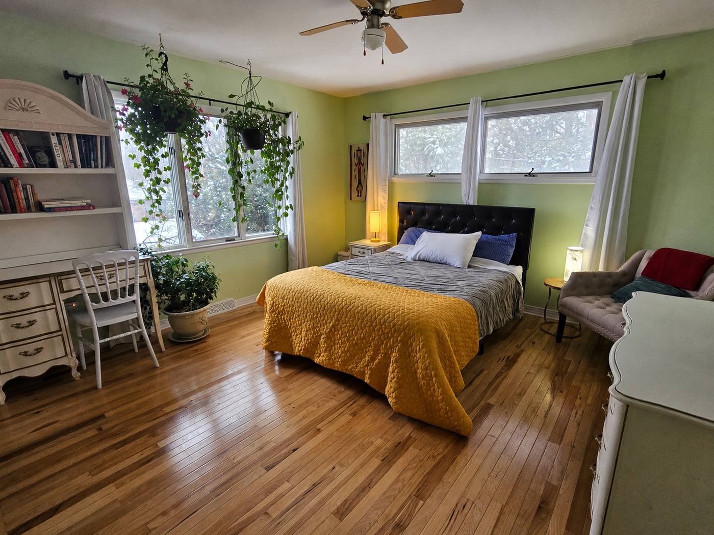 A bedroom in soft green with trailing plants above a built-in desk, wide windows and a warm oak floor.