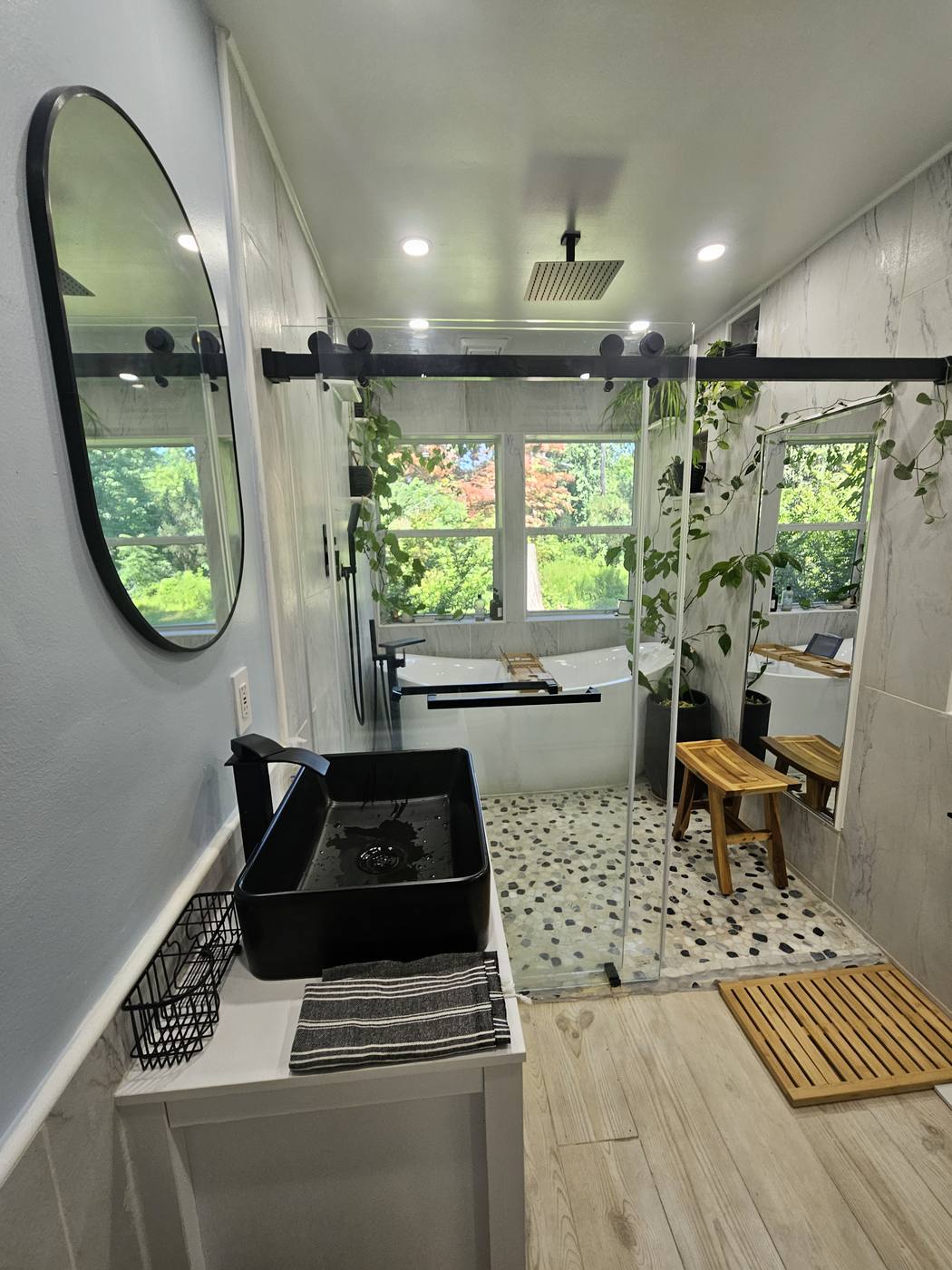 A bathroom with a black vessel tub, pebble-tiled floor, teak bench and plants on the windowsill.