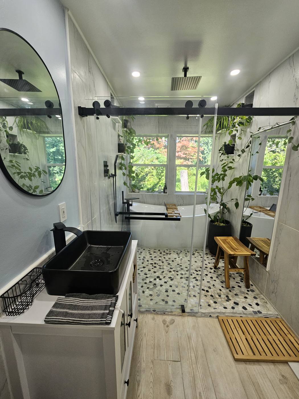 A bathroom vanity with a round black mirror, marble surround and a window looking into greenery.